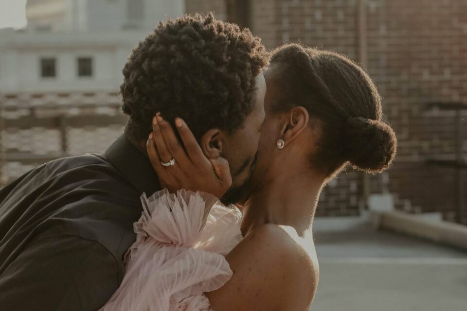 139997 a man kisses a woman on the cheek on a roof in a city the woman is wearing a pink chiffon off the shoulder dress unsplash kadariu seegars
